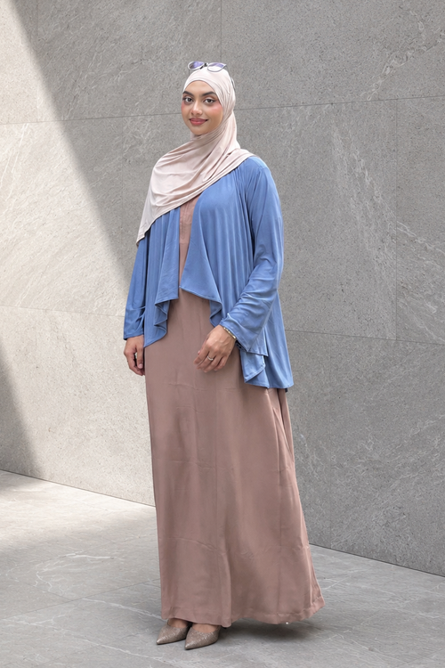 Woman wearing a blue lily shrug and beige hijab standing against a gray wall.