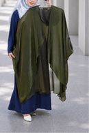 A woman wearing a navy blue dress and light blue hijab holds up a large olive green sheer shawl or wrap over her arm in a modern architectural corridor with columns.