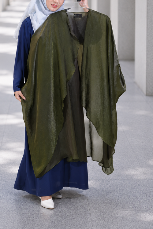 A woman wearing a navy blue dress and light blue hijab holds up a large olive green sheer shawl or wrap over her arm in a modern architectural corridor with columns.