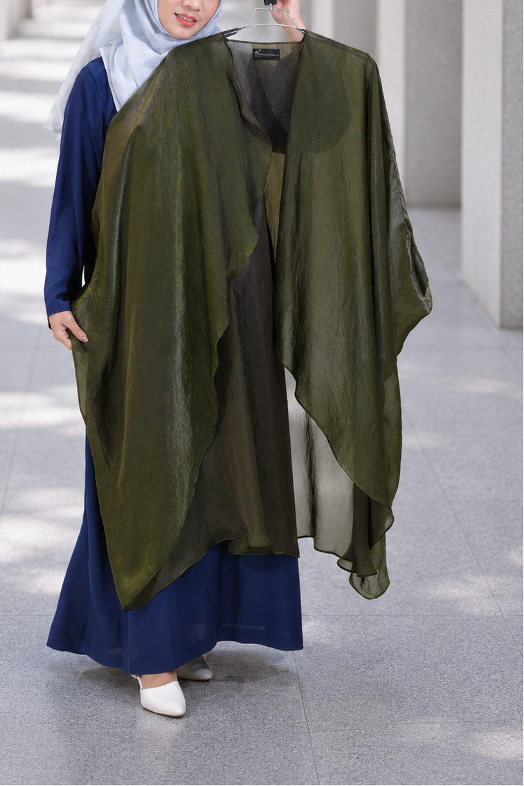 A woman wearing a navy blue dress and light blue hijab holds up a large olive green sheer shawl or wrap over her arm in a modern architectural corridor with columns.
