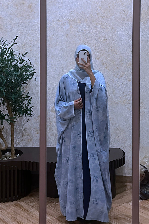 Instagram influencer is wearing a light blue shrug of modest collection taking a mirror selfie in a room with a plant and door.