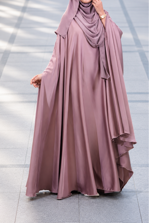 A woman wearing a flowing mauve-colored abaya and matching hijab, standing on a tiled outdoor walkway.