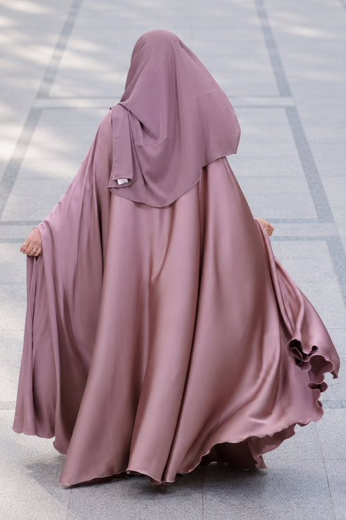 A woman wearing a flowing mauve-colored modest garment with a matching head covering, photographed from behind in an outdoor paved walkway.