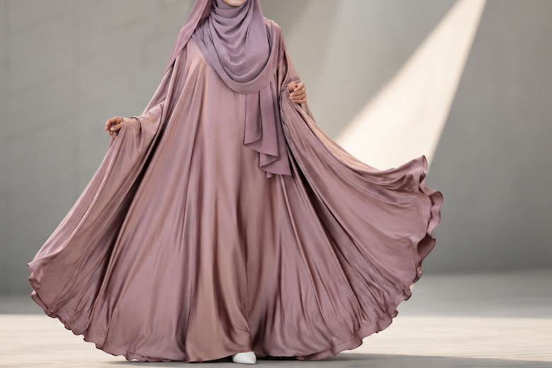 Woman in a flowing mauve dress and hijab standing in a modern architectural setting.