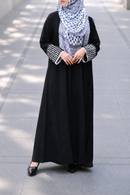 A woman wearing a black keffiyeh  abaya with patterned long sleeves and a black-and-white checkered hijab, standing on a paved outdoor walkway.
