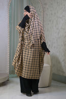 A woman wearing a beige and black plaid hijab and a matching long wrap dress with black sleeves and wide-leg black pants, standing in a modern interior with arched mirrors.