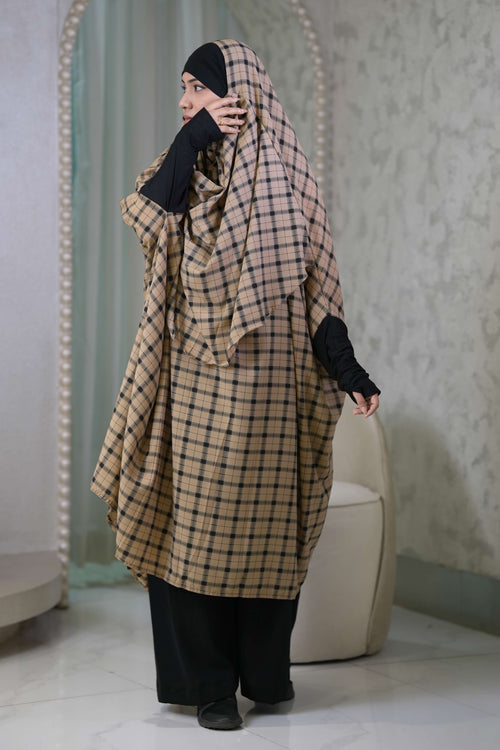 A woman wearing a beige and black plaid hijab and a matching long wrap dress with black sleeves and wide-leg black pants, standing in a modern interior with arched mirrors.