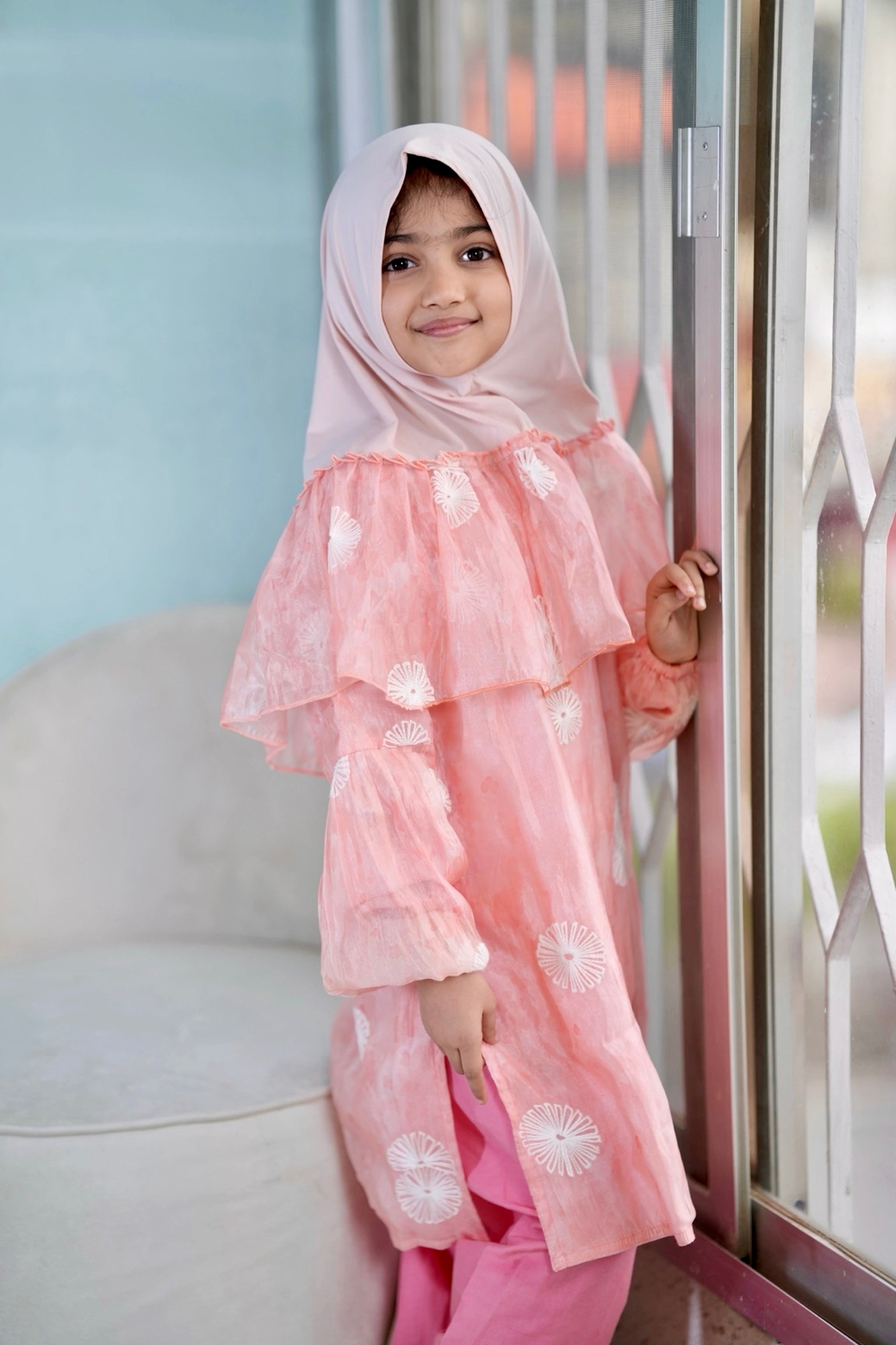 A young girl wearing a light pink hijab and a matching pink long-sleeve tunic with white floral embroidery, standing indoors next to a glass door.