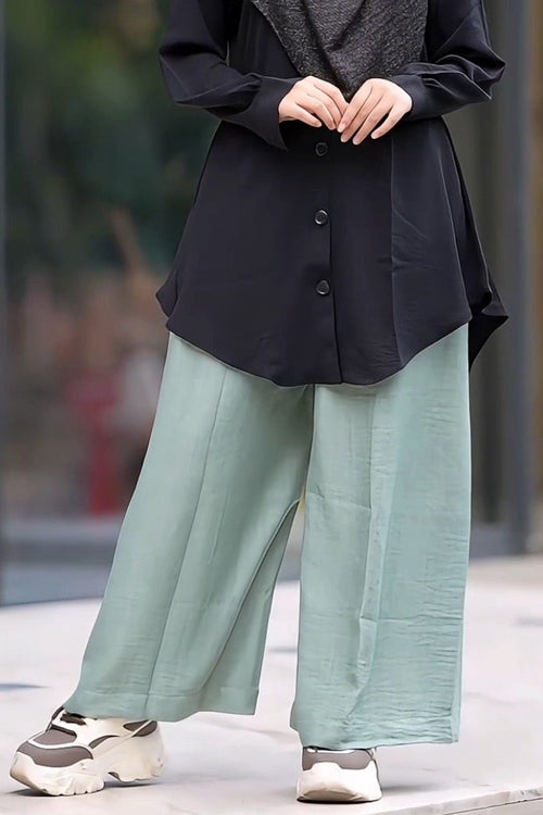 A woman wearing a black button-up tunic, wide-leg sage green maternity formal pants, and a dark gray patterned hijab stands outdoors in front of a modern glass building.