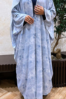 Person wearing a light blue floral abaya in an indoor setting with a plant in the background.