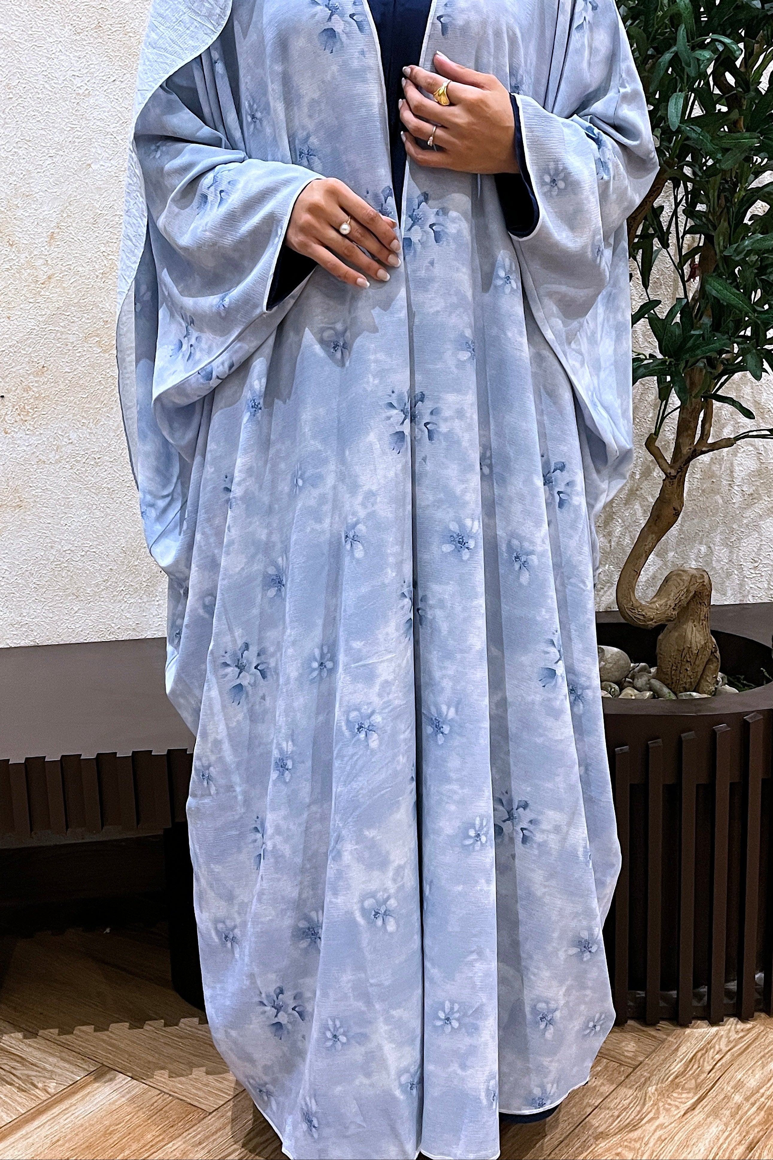 Person wearing a light blue floral abaya in an indoor setting with a plant in the background.