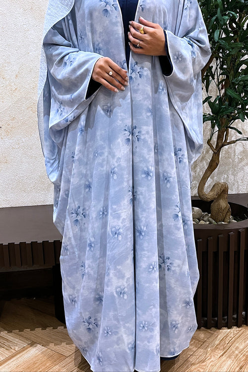 Person wearing a light blue floral abaya in an indoor setting with a plant in the background.