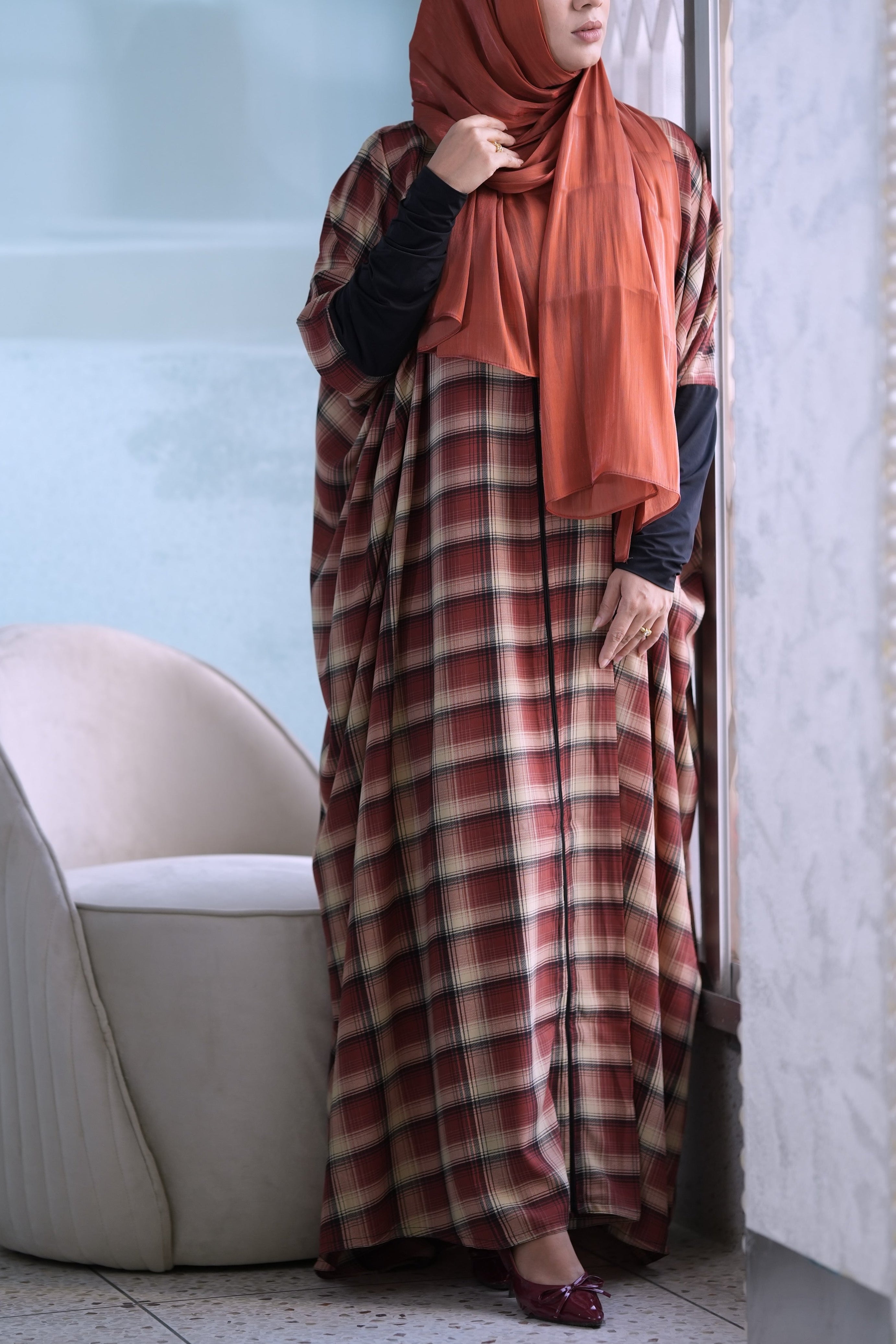 A woman wearing a rust-orange hijab and a long red and brown safia plaid kaftan abaya with black long sleeves, standing in a doorway in front of a light blue wall.