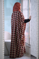 A woman wearing a rust-red hijab and a matching long plaid shawl over a black long-sleeve top stands by a window in a bright indoor setting.