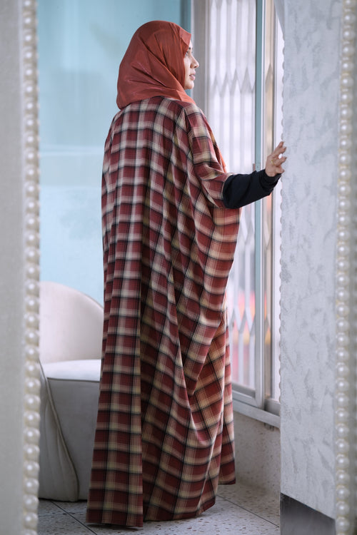 A woman wearing a rust-red hijab and a matching long plaid shawl over a black long-sleeve top stands by a window in a bright indoor setting.