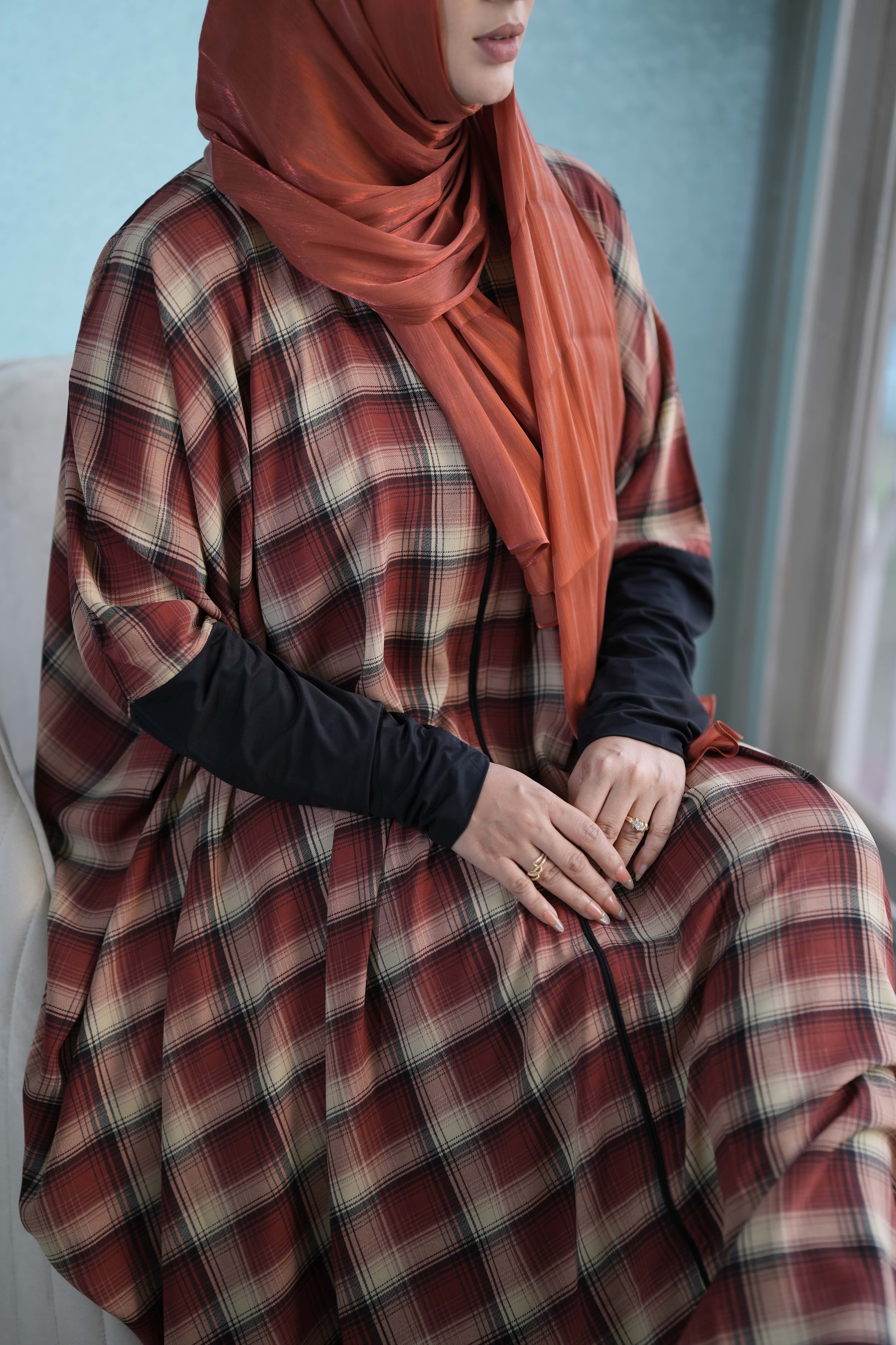 A woman wearing a rust-orange hijab and a matching rust-red and black Safiya plaid Kaftan abaya with black long-sleeve undersleeves, seated indoors.