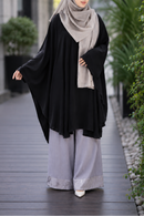 A woman wearing a black short kaftan, light gray wide-leg pants, and a light gray hijab walks along a paved outdoor walkway.