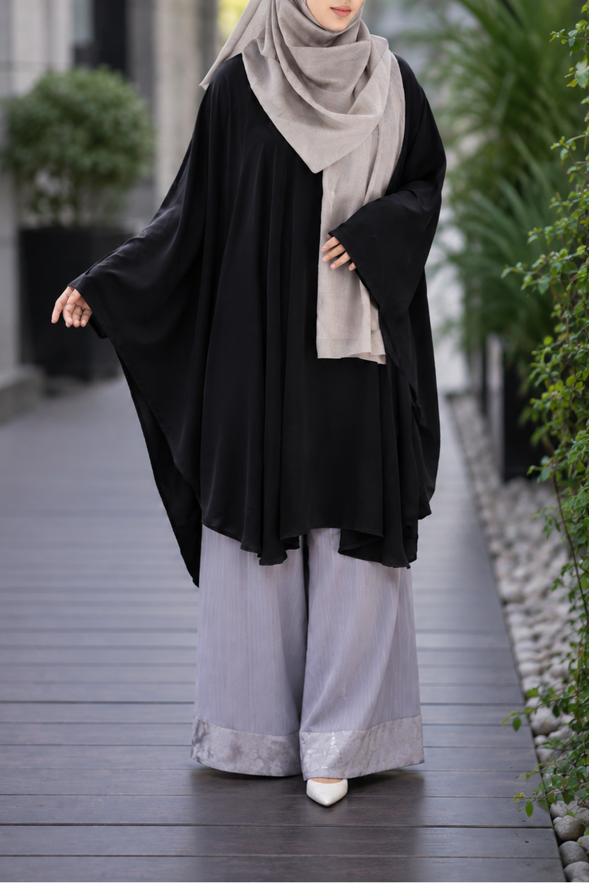 A woman wearing a black short kaftan, light gray wide-leg pants, and a light gray hijab walks along a paved outdoor walkway.