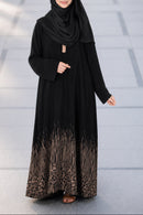 A woman wearing a black abaya with a brown gradient lace hem and a matching black hijab, standing on a paved outdoor walkway.
