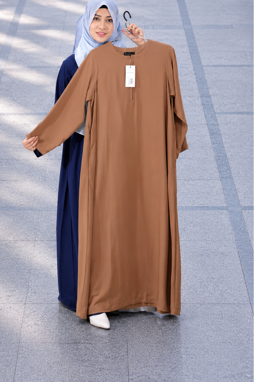 A woman wearing a light blue hijab and navy abaya stands outdoors holding up a brown long-sleeve abaya dress on a hanger in front of a gray tiled walkway.