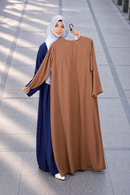 A woman wearing a navy blue abaya and light blue hijab stands outdoors holding up a brown abaya on a hanger to show the color and fit. The background is a clean, modern tiled walkway.