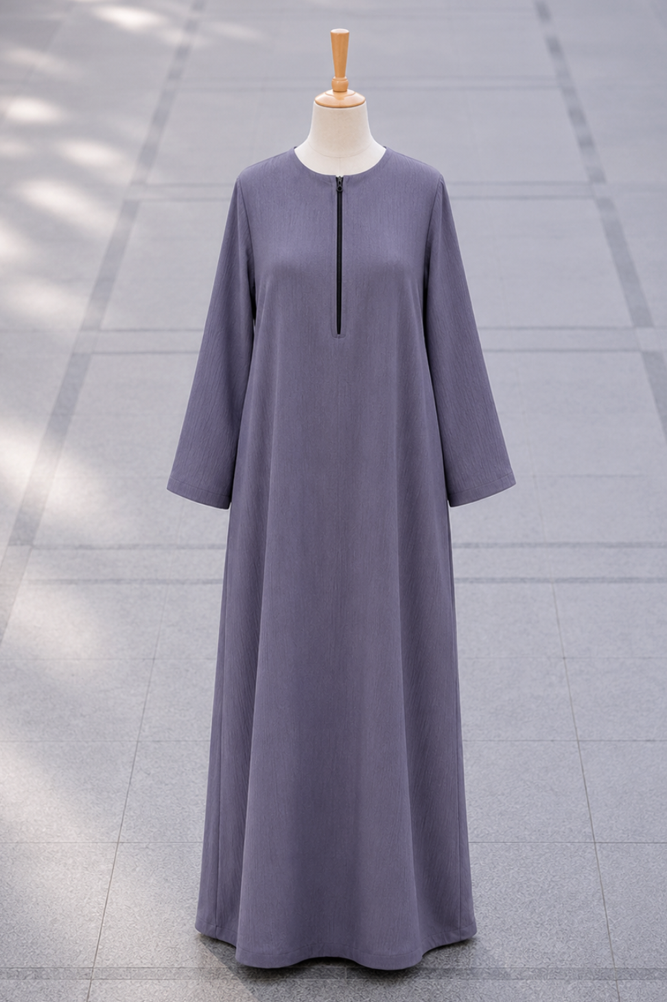 A long-sleeved, floor-length abaya dress in a muted dusty purple-gray color, displayed on a mannequin against a gray tiled outdoor background.