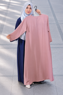 A woman wearing a navy blue abaya and a light blue hijab holds up a dusty pink abaya on a black hanger in front of her. She is standing on a gray tiled outdoor walkway.