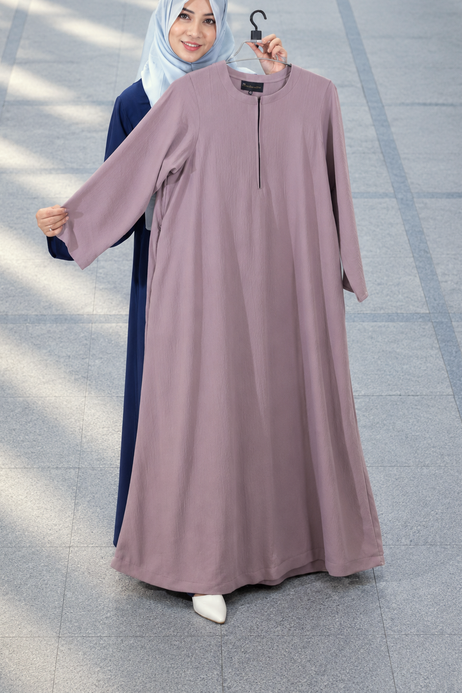 A woman wearing a light blue hijab and a dark navy abaya holds up a mauve-colored abaya on a hanger in front of a tiled outdoor walkway.