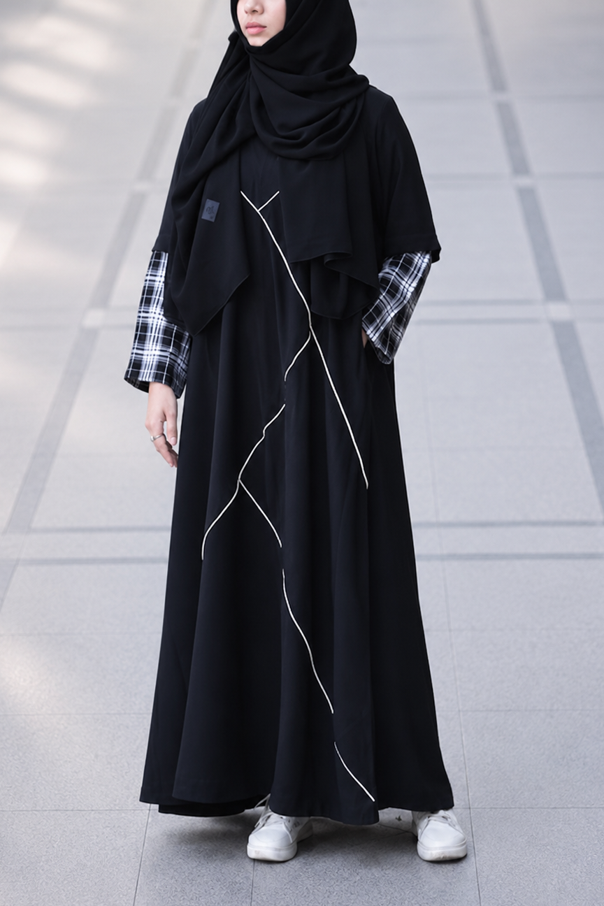 A woman wearing a long black abaya dress with white geometric line details, a black hijab, and a black-and-white plaid shirt underneath, standing on a gray tiled floor.
