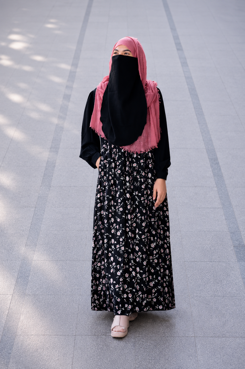 A woman wearing a black long-sleeve maxi dress with a small white floral print, a black face veil, and a pink hijab, standing on a gray tiled outdoor walkway.