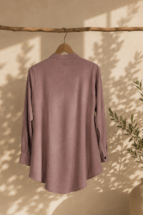 Mauve shirt hanging on a wooden hanger against a beige wall with a plant in the corner.