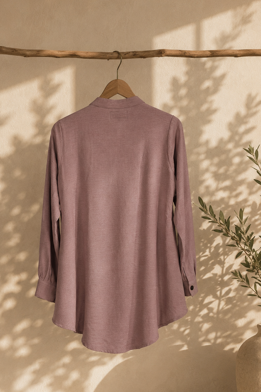 Mauve shirt hanging on a wooden hanger against a beige wall with a plant in the corner.