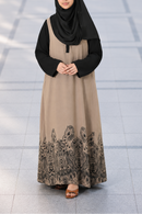 A woman wearing a black hijab and a long beige abaya of Modest Collection with a black printed border, standing on a tiled floor.